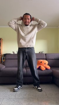 Um moletom bege com um bolso frontal, jeans cinzentos e ténis preto e branco. Um brinquedo de polvo de pelúcia laranja está sobre um sofá cinza escuro.