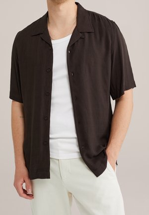 Mann trägt offene schwarze kurzärmelige Knopfleiste über weißem T-Shirt und hellen Hosen, eine Hand in der Tasche.