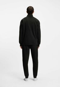 Tuta atletica nera con colletto con zip, pannello superiore testurizzato e pantaloni affusolati con polsini elasticizzati, vista da dietro.