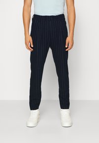 Man som bär marinblå randiga byxor och vita sneakers, stående mot en enkel vit bakgrund.