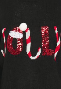 Pull en tricot noir avec le mot "Jolly" en sequins rouges, agrémenté d'un pompon blanc et de motifs de cannes de Noël rayées.