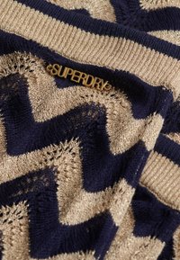 Marineblå og guld strikket tekstil med et zigzag-mønster, med en struktureret overflade og en guld logo-accents der læser "SUPERDRY."