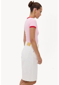 Femme aux cheveux en chignon, portant des boucles d'oreilles créoles dorées, un t-shirt rose à manches courtes avec bordure rouge, et une jupe blanche jusqu'aux genoux, vue de profil.