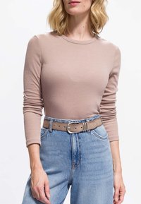 Frau in einem beigen, gerippten Langarmshirt, das in eine hochgeschnittene hellblaue Jeans gesteckt ist, mit einem taupefarbenen Wildledergürtel und einer silbernen Schnalle.