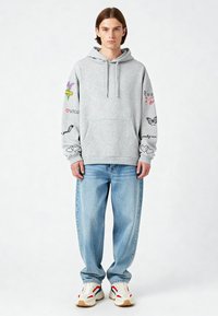 Sudadera gris con bolsillo frontal, gráficos y texto coloridos en contraste en las mangas, combinada con vaqueros claros y sueltos de color azul y zapatillas blancas.