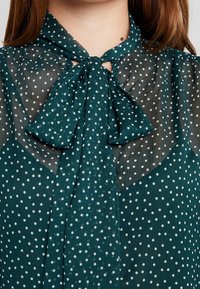 Blusa trasparente color teal con pois bianchi. Presenta un colletto a fiocco e una scollatura a chiave. Tessuto morbido con leggera trasparenza e design fluido.