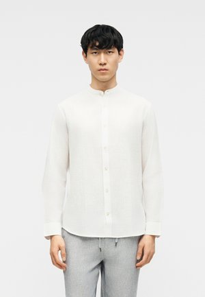 Hombre vistiendo una camisa blanca de manga larga con cuello mandarín y pantalones gris claro, de pie contra un fondo blanco liso.