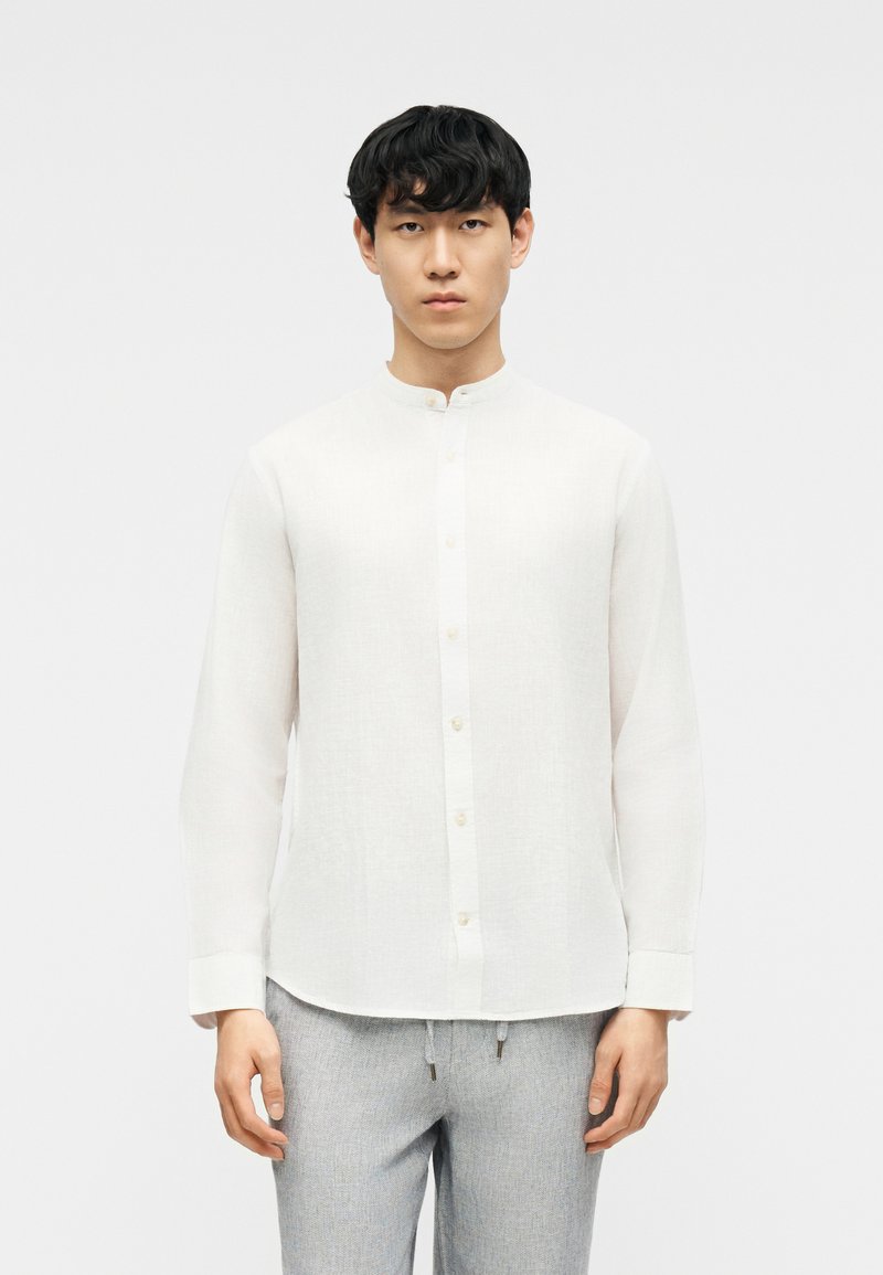 Άνδρας που φοράει λευκό πουκάμισο με γιακά μανταρίνι και μακριά μανίκια, καθώς και ανοιχτόχρωμο γκρι παντελόνι, στέκεται μπροστά σε λευκό απλό φόντο.