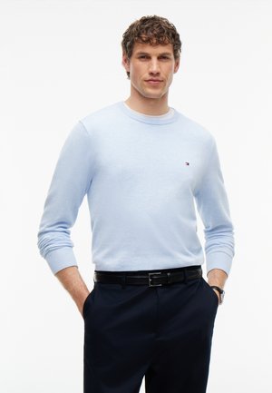 Mand med krøllet hår iført en lyseblå sweater og mørke bukser, hænder i lommerne, stående mod en ensfarvet hvid baggrund.