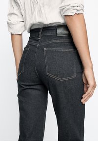 Dark denim jeans featuring a fitted cut, visible back pockets, and contrasting stitching. The waistband has a black leather label accent.