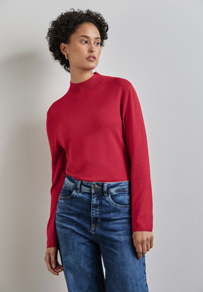 Rotes Strickoberteil mit hohem Kragen und langen Ärmeln, kombiniert mit verwaschenen blauen Jeans im Five-Pocket-Design mit Knopf.