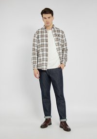 Koszula w kratę z długim rękawem w odcieniach beżowym i niebieskim na białym T-shircie, zestawiona z ciemnoniebieskimi dżinsami i brązowymi butami.