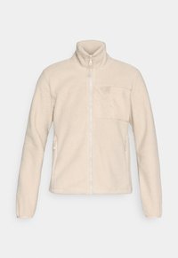 Chaqueta de forro polar beige con cuello alto, cremallera frontal completa y un bolsillo en el pecho. Cuenta con bolsillos laterales y una superficie texturizada y suave.