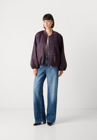 Giacca bomber bordeaux con collo rotondo, zip frontale e due tasche laterali, abbinata a jeans blu a gamba larga e scarpe nere con punta.