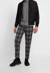 Homme portant un pull à col roulé gris en maille torsadée, une veste bomber noire, un pantalon à carreaux gris et des baskets noires, debout devant un fond blanc.