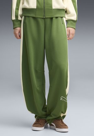 Person iført grønne og cremefarvede trackpants med brede ben og sidestriber, matchende jakke og brune sneakers, står mod en ensfarvet baggrund.