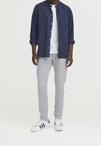 Camicia blu navy abbottonata, t-shirt bianca sotto, pantaloni grigi e sneaker bianche con strisce nere. Outfit casual con maniche della camicia arrotolate.