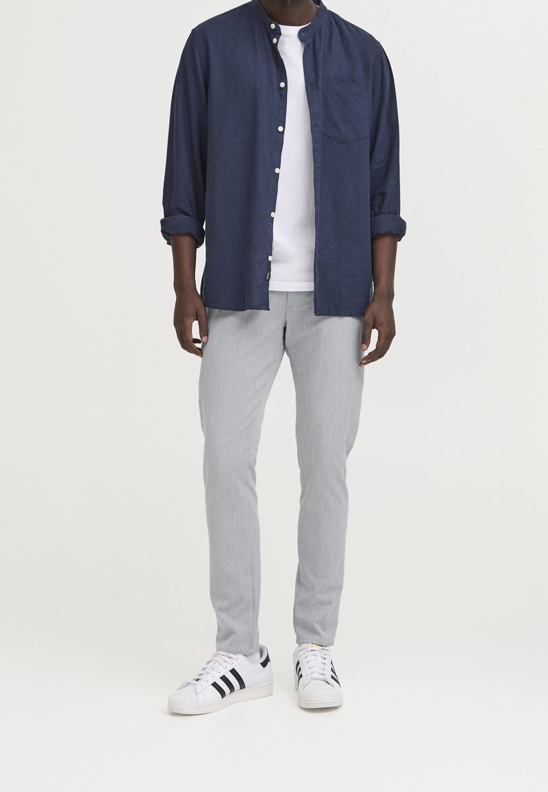 Camicia blu navy abbottonata, t-shirt bianca sotto, pantaloni grigi e sneaker bianche con strisce nere. Outfit casual con maniche della camicia arrotolate.