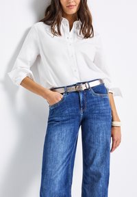 Bíla košile s knoflíky a ohrnutými rukávy, spojená s modrými denimovými širokými kalhotami a metalickou stříbrnou páskem s hladkým povrchem.