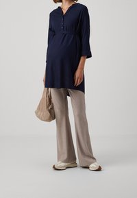 Navy blue maternity tunic with a collar and front buttons, paired with beige ribbed wide-leg trousers and light-colored sneakers.