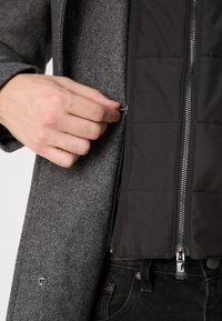 Main fermant une petite fermeture éclair sur un manteau en laine grise superposé à une veste matelassée noire avec une fermeture éclair centrale et un pantalon noir.
