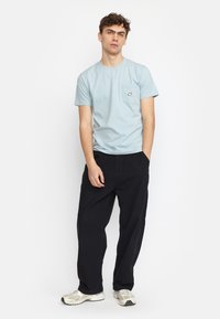 Lichtblauw katoenen T-shirt met een zak, voorzien van een geborduurd decoratief patch, gecombineerd met losse zwarte broeken en grijze sneakers.