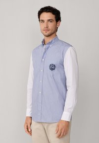 Uomo con una camicia button-up a righe blu e bianche con maniche lunghe bianche e pantaloni beige, in piedi di fronte a uno sfondo neutro.
