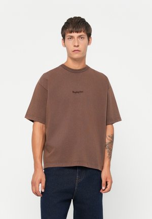 Young man with short brown hair wearing a brown oversized t-shirt and dark blue jeans, standing against a plain white background.