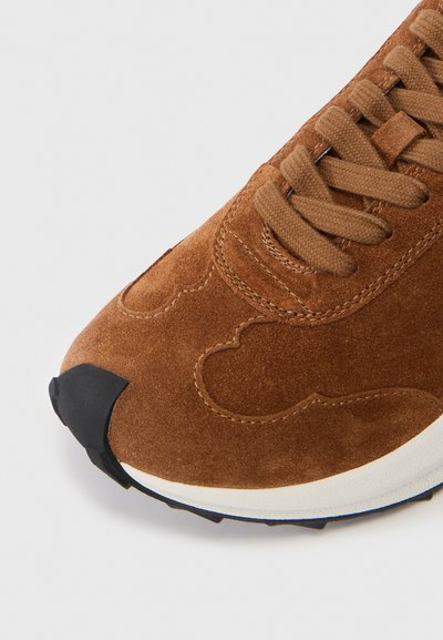Adidași din suede maro cu șireturi asortate, talpă intermediară albă și vârf și talpă din cauciuc negru, prezentați dintr-un unghi frontal superior.