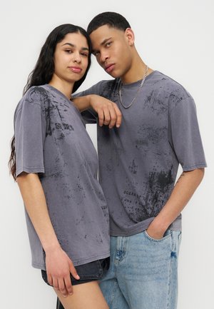 Junge Frau und junger Mann tragen passende graue, abstrakt gemusterte, übergroße T-Shirts und posieren vor einem schlichten Hintergrund.