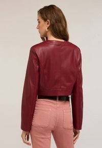 Giacca in pelle bordeaux tagliata con maniche lunghe, finitura liscia e scollatura rotonda, abbinata a pantaloni aderenti rosa pallido.