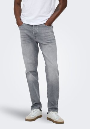 Man draagt lichtgrijze rechte jeans, witte sneakers en een wit t-shirt met handen in de zakken, staand op een effen achtergrond.