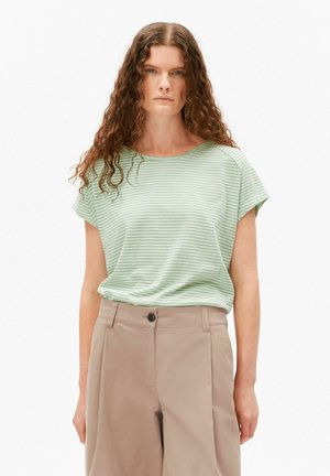 Woman with long wavy hair wearing a light green and white striped short-sleeve shirt and beige high-waisted pants against a white background.