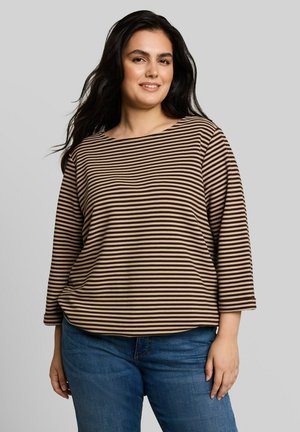Femme aux longs cheveux foncés portant un haut à manches longues rayé marron et beige et un jean bleu, debout devant un fond uni.