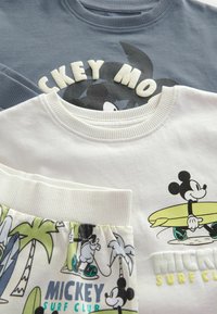 Tre capi di abbigliamento: un felpa blu con una grafica di "Topolino", una maglietta bianca con Topolino che tiene una tavola da surf e dei pantaloni corti a fantasia.