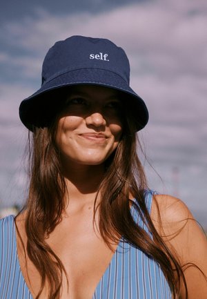 Young woman with long brown hair wearing a blue striped top and navy bucket hat with "self." text, smiling against cloudy sky background.