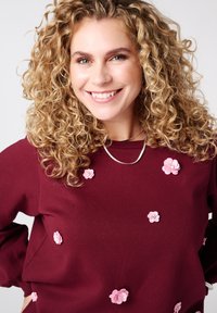 Glimlachende vrouw met krullend blond haar, gekleed in een bordeauxrood sweater versierd met roze 3D bloemapplicaties en een zilveren ketting.