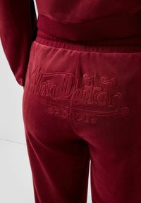 Close-up van bordeauxrode velours broeken met elastische tailleband en het reliëf Von Dutch Originals logo op het achterzakgebied.