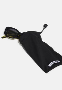 Black sunglasses with a square frame partially inside a black fabric pouch. Pouch has a drawstring and "ARNETTE" label.