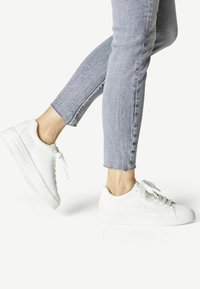 Weiße Sneaker aus glattem Material, mit runder Zehenpartie, flacher Sohle und strukturiertem Schnürdesign, kombiniert mit leicht verwaschenen Jeans.