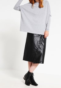 Graues, oversized Langarmoberteil kombiniert mit einem schwarzen Leder-Midi-Rock mit diagonaler Schlitze und knöchelhohen schwarzen Stiefeln.