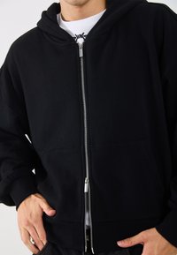 Sudadera con cremallera negra y capucha amplia, fabricada en un tejido suave. Cuenta con una cremallera de metal plateado y dos bolsillos laterales. Ajuste texturizado y casual.