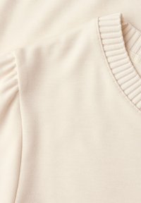 Jasnobeżowy dzianinowy top z dekoltem w serek, z prążkowanym obszyciem przy dekolcie i plisami na ramionach, wykonany z miękkiego materiału.