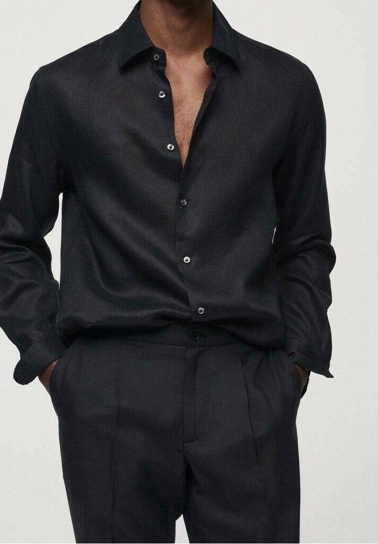 Homme portant une chemise noire à boutons dont trois sont déboutonnés et un pantalon noir taille haute, les mains dans les poches.
