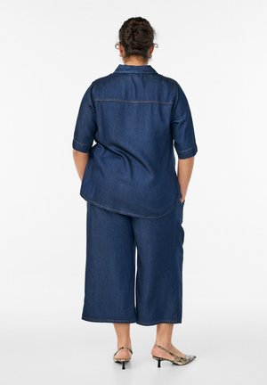 Frau mit geflochtenem Haar, die ein dunkelblaues Jeanshemd und weit geschnittene, knöchellange Hosen trägt, steht mit den Händen in den Taschen und ist von hinten zu sehen.