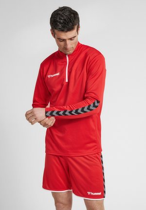 Mann passt den Ärmel eines roten Sporttops mit schwarzen Chevron-Streifen an, trägt passende rote Shorts mit weißen Hummel-Logos.