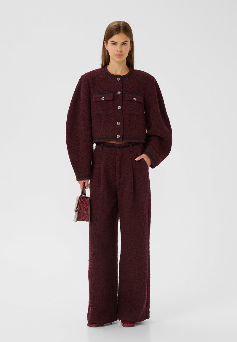 Burgunderrotes, strukturiertes zweiteiliges Outfit mit kurzer Jacke, zwei Taschen und schwarzem Besatz, kombiniert mit weit geschnittenen Hosen und einer kleinen gemusterten Handtasche.