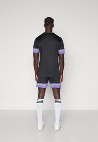 Maillot de sport noir avec des rayures violettes et vertes sur les manches, associé à un short noir avec des accents similaires, porté avec des chaussettes et des chaussures blanches.