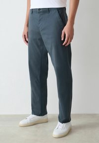 Mann in dunkelblauen geraden Hosen und weißen Sneakers steht auf einem hellgrauen Boden vor einer weißen Wand.