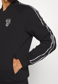 Giacca nera con zip e colletto alto, caratterizzata da strisce bianche lungo le maniche e un piccolo logo grafico sul petto.